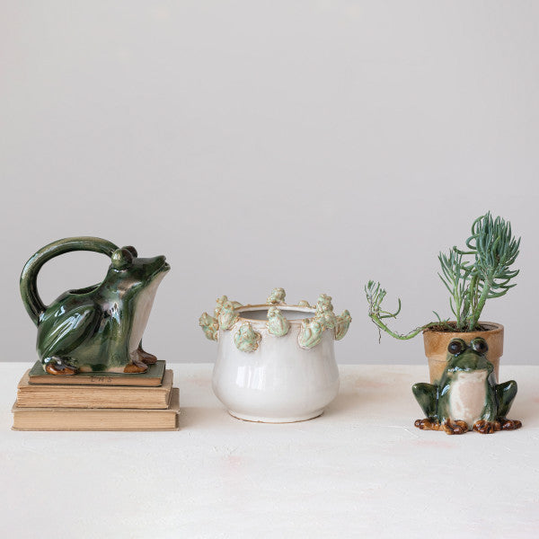 A stoneware frog planter with a round body and a smiling face, primarily green with white and brown details.